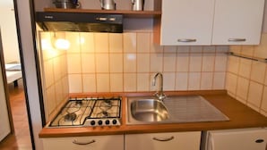 Une cuisine compacte équipée d’une cuisinière à gaz, d’un évier et d’un petit réfrigérateur. On y trouve des placards au-dessus de la cuisinière et une étagère contenant des ustensiles de cuisine.