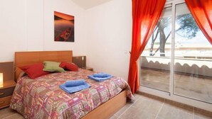 Un dormitorio con cama, almohadas, una mesita de noche, lámpara, ventana con cortinas y un cuadro en la pared.