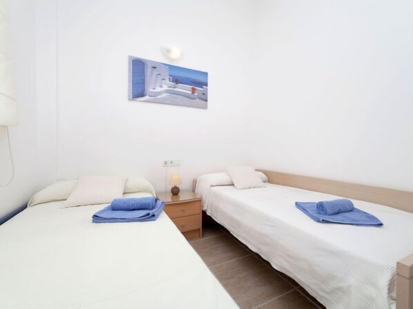 A hotel room with two single beds, a nightstand with a lamp, and a wall-mounted picture of a coastal scene. - Vacation home Juanes by Interhome (L'Escala)