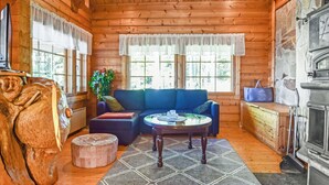 A cozy cabin interior with a wooden ceiling, a stone fireplace, a blue sofa, a round coffee table, and a large window with curtains. - Linnunlaulu by Interhome (Lapinlahti)
