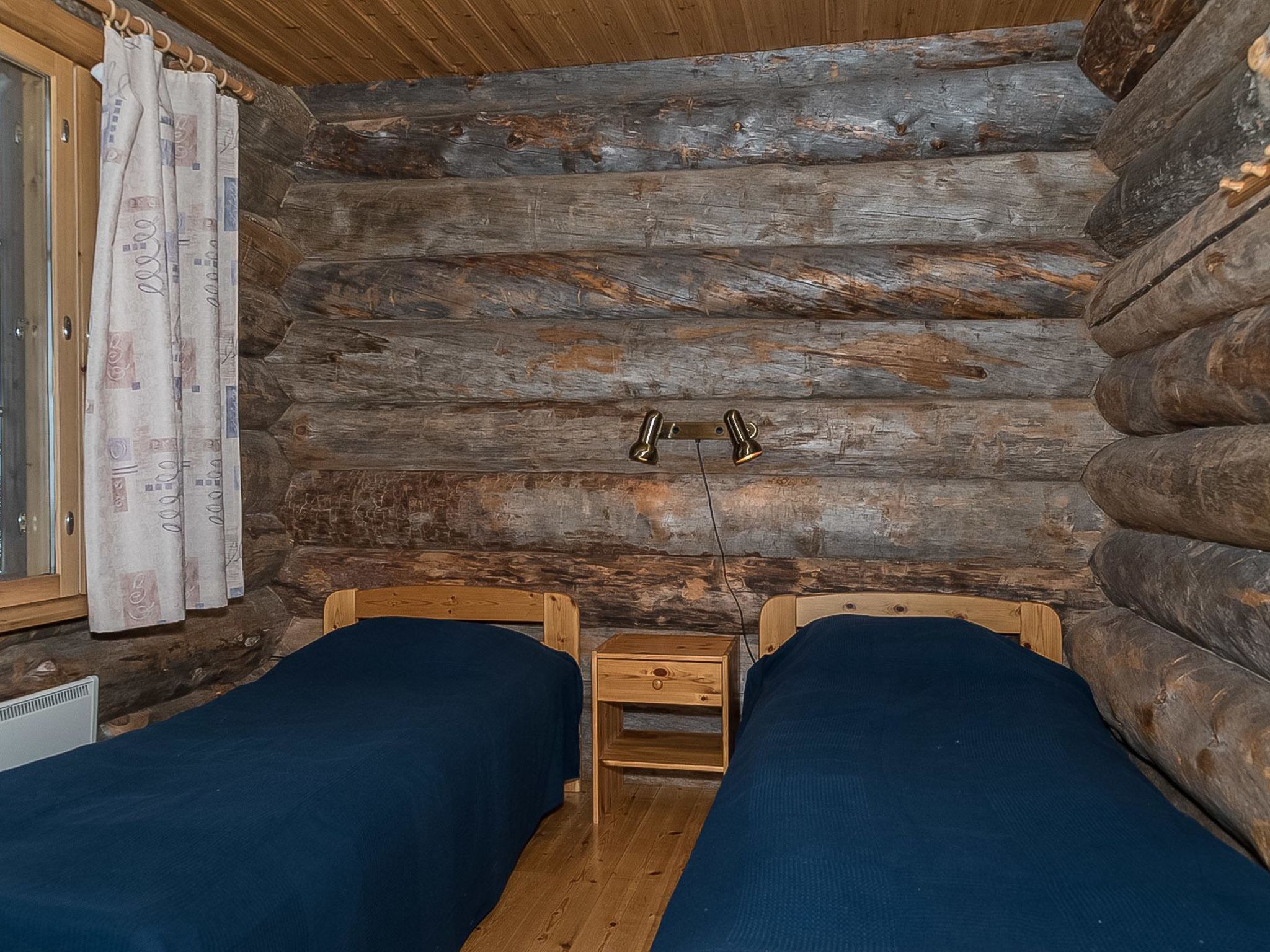 Una habitación tipo cabaña de troncos con dos camas de madera, una mesita de noche de madera y una ventana con cortinas.