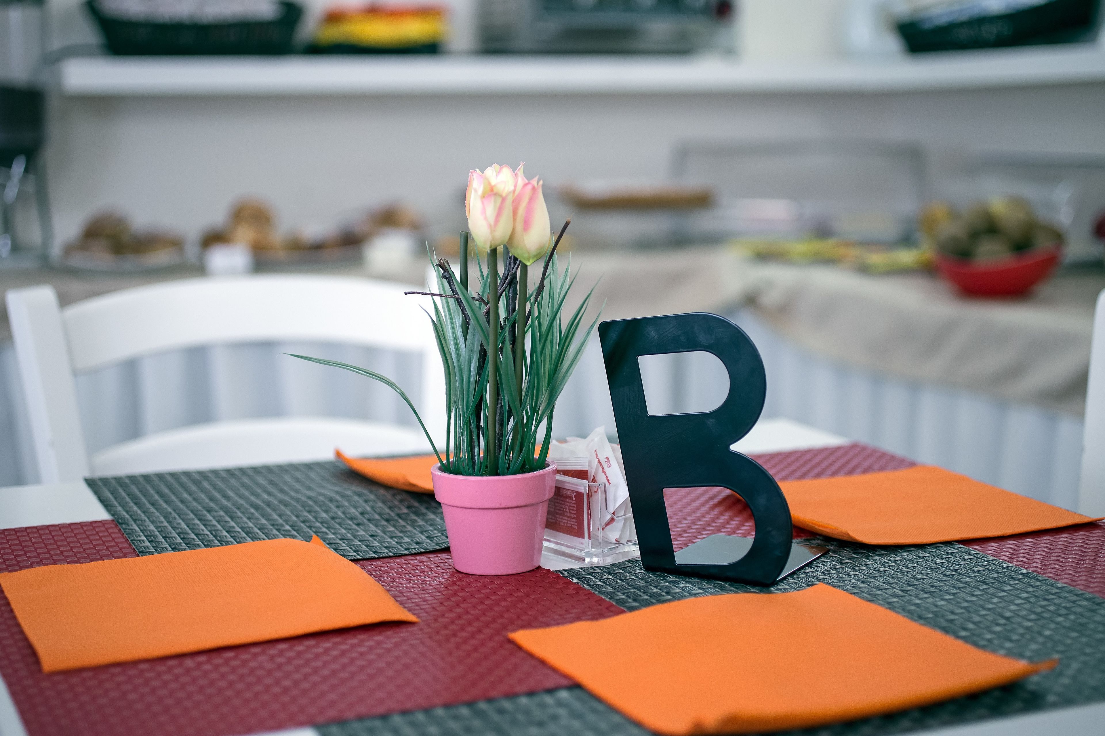 Una mesa con una maceta rosa, una letra 'B' negra y manteles individuales naranja.