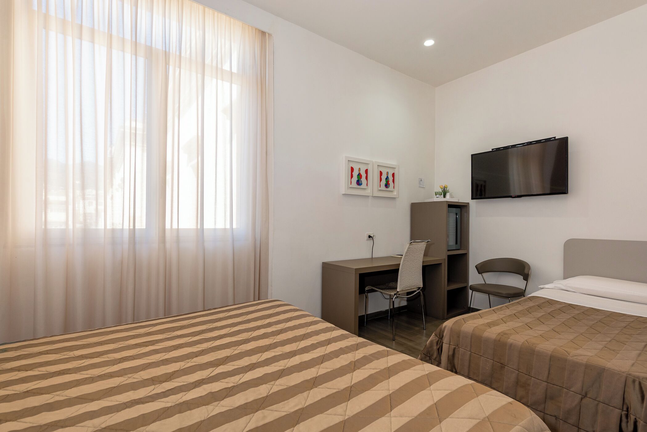 A hotel room with two beds, a desk, a chair, a TV, and a window with sheer curtains.