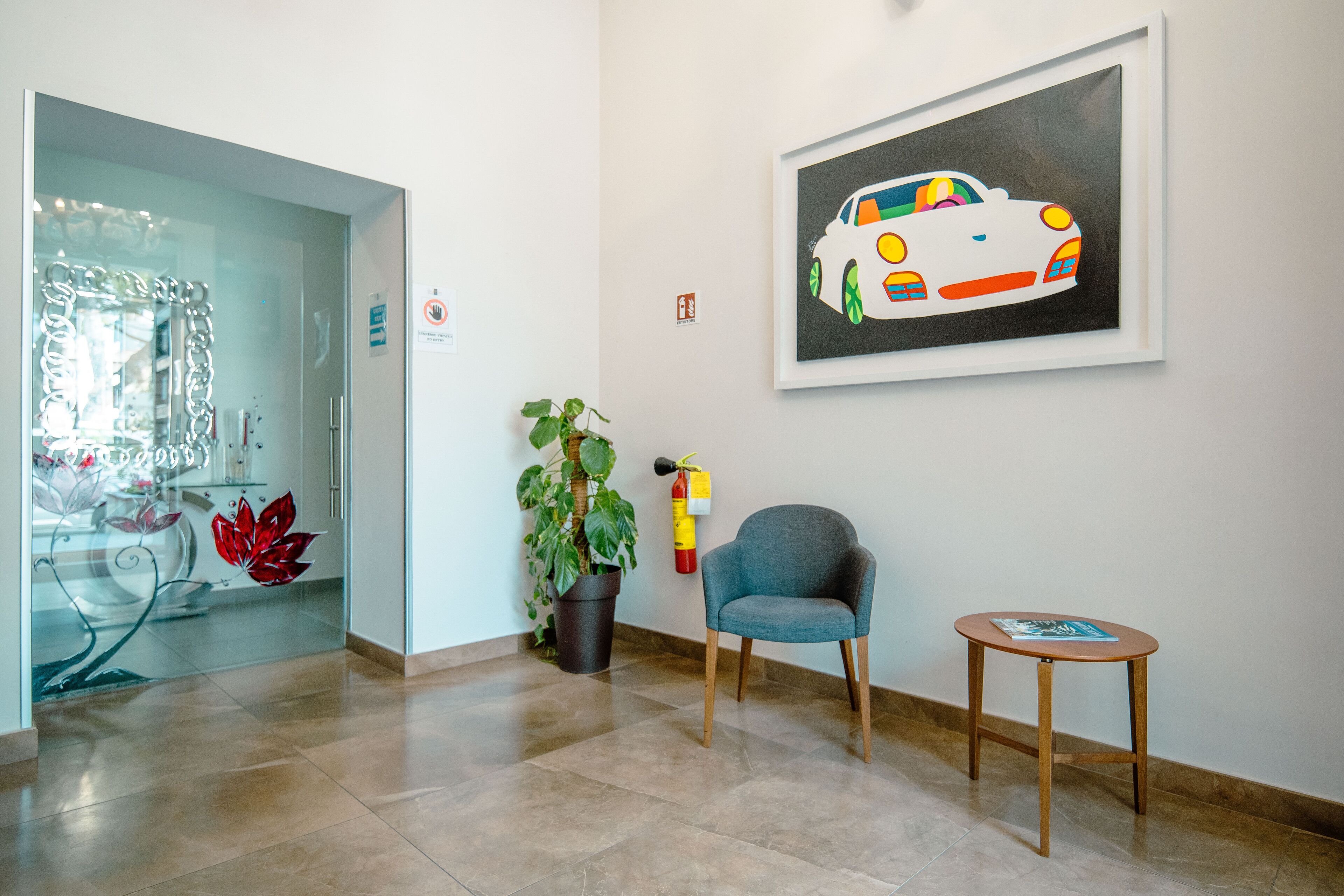 Un vestíbulo moderno con una puerta de cristal, una planta en maceta, un sillón azul, una pequeña mesa de madera y una obra de arte enmarcada de un coche.