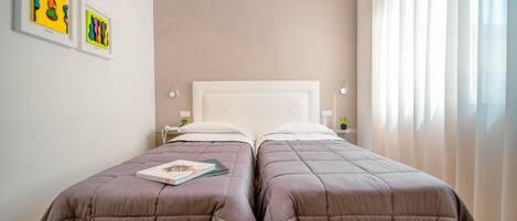 Cama doble con cabecera, dos mesitas de noche y ventana con cortinas.