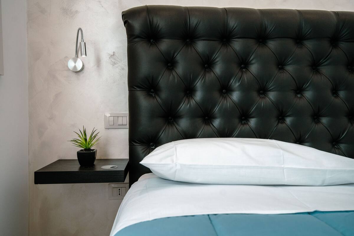 Een slaapkamer met een gestoffeerd hoofdeinde, een nachtkastje met een plant en een wit kussen.