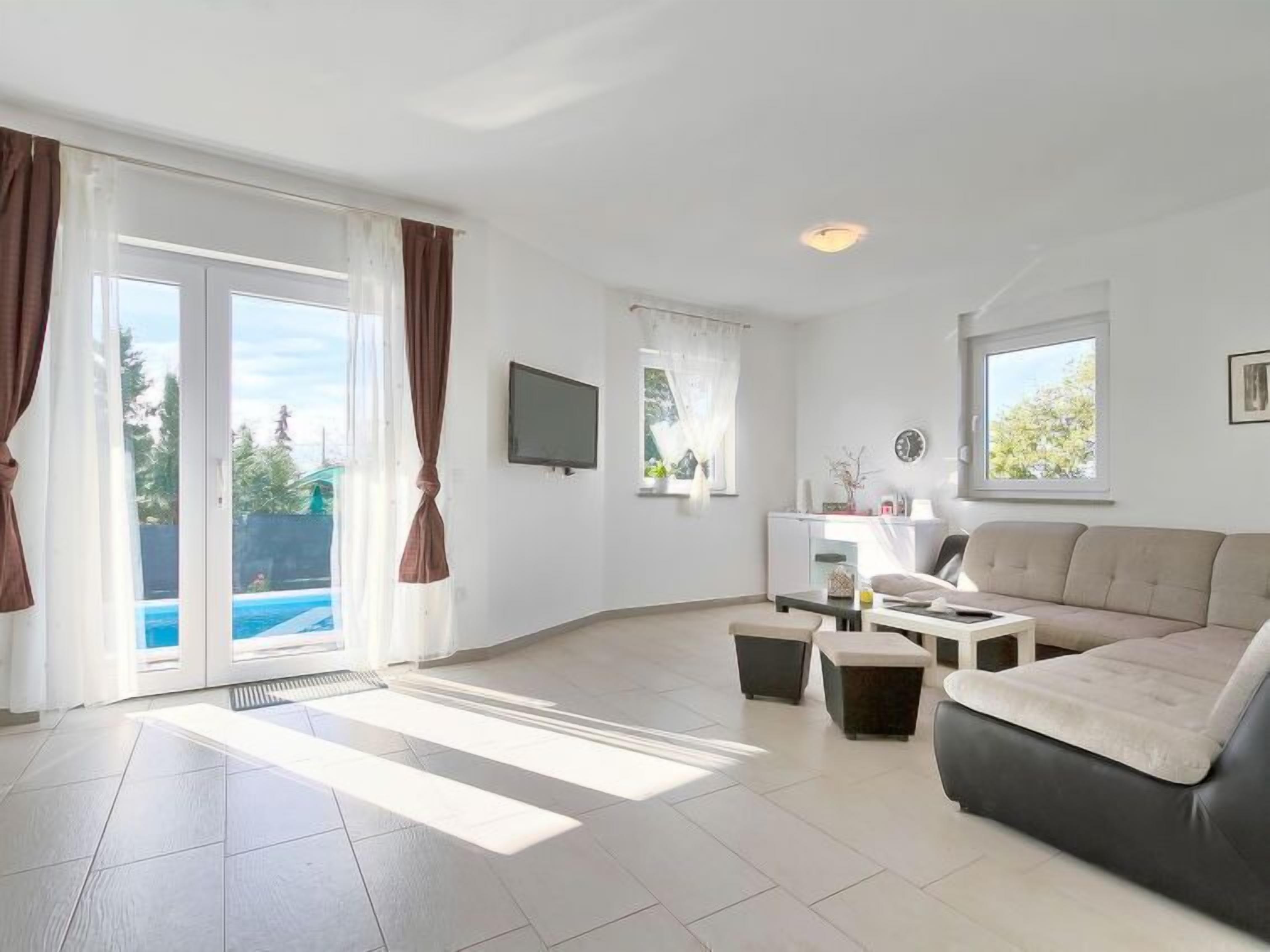 Amplia sala de estar con sofá, una mesa de centro y televisor. Cuenta con ventanas grandes con cortinas, una puerta corrediza de vidrio hacia una piscina y un escritorio pequeño con silla.