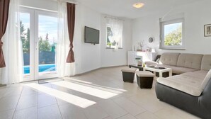 Un salon spacieux comprenant un canapé, une table basse et un téléviseur. De grandes fenêtres sont présentes, ainsi que des rideaux, une porte-fenêtre coulissante donnant sur une piscine et un petit bureau avec une chaise.