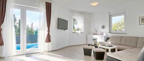 Amplia sala de estar con sofá, una mesa de centro y televisor. Cuenta con ventanas grandes con cortinas, una puerta corrediza de vidrio hacia una piscina y un escritorio pequeño con silla.