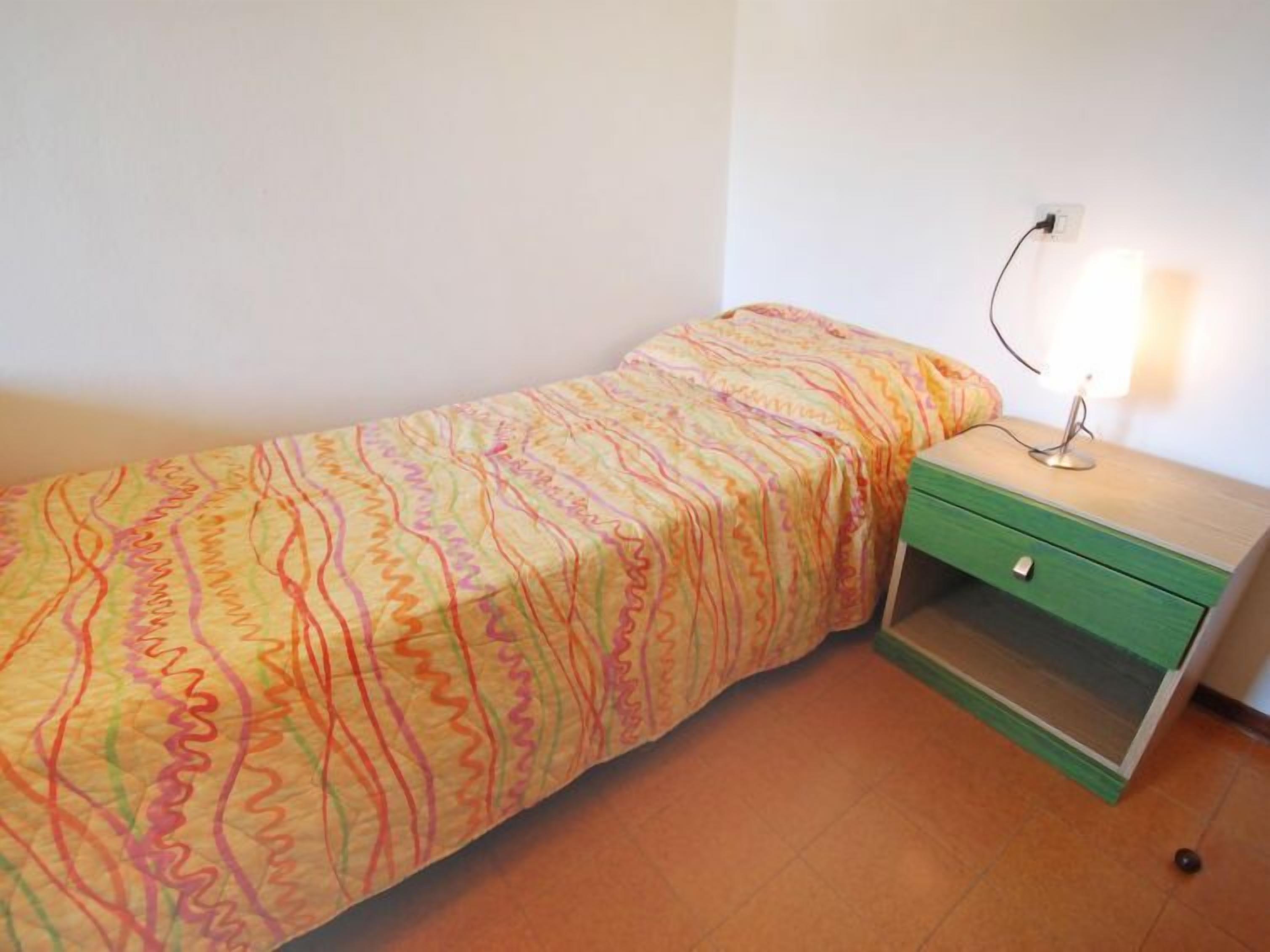 A small, single-bed room with a colorful bedspread, a bedside table with a lamp, and a green drawer unit.