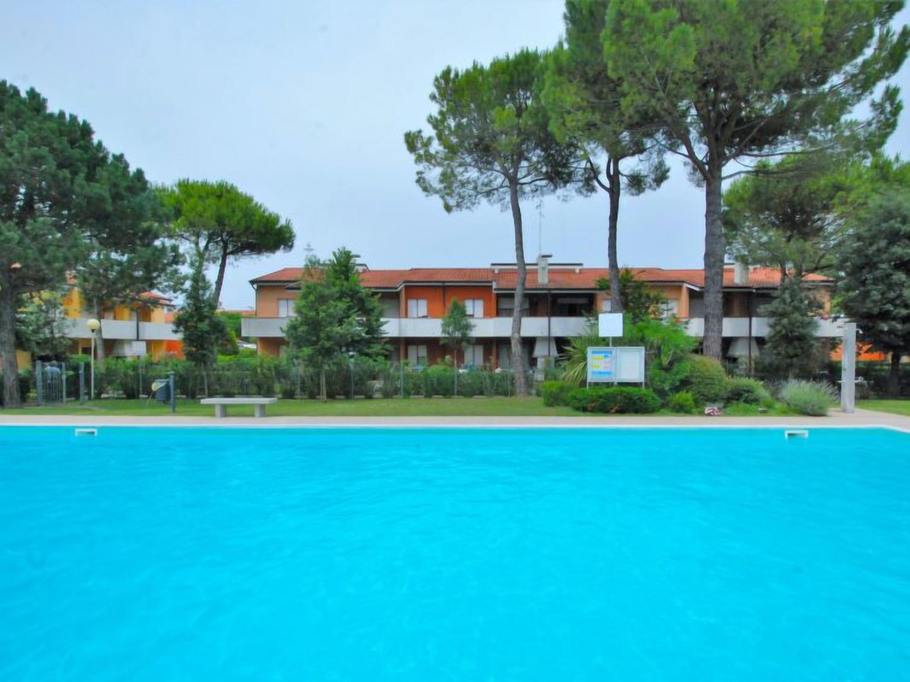 A swimming pool with a clear blue water surface, surrounded by a well-maintained lawn and trees. In the background, there are multi-story buildings with orange and white facades, and a signboard is visible.