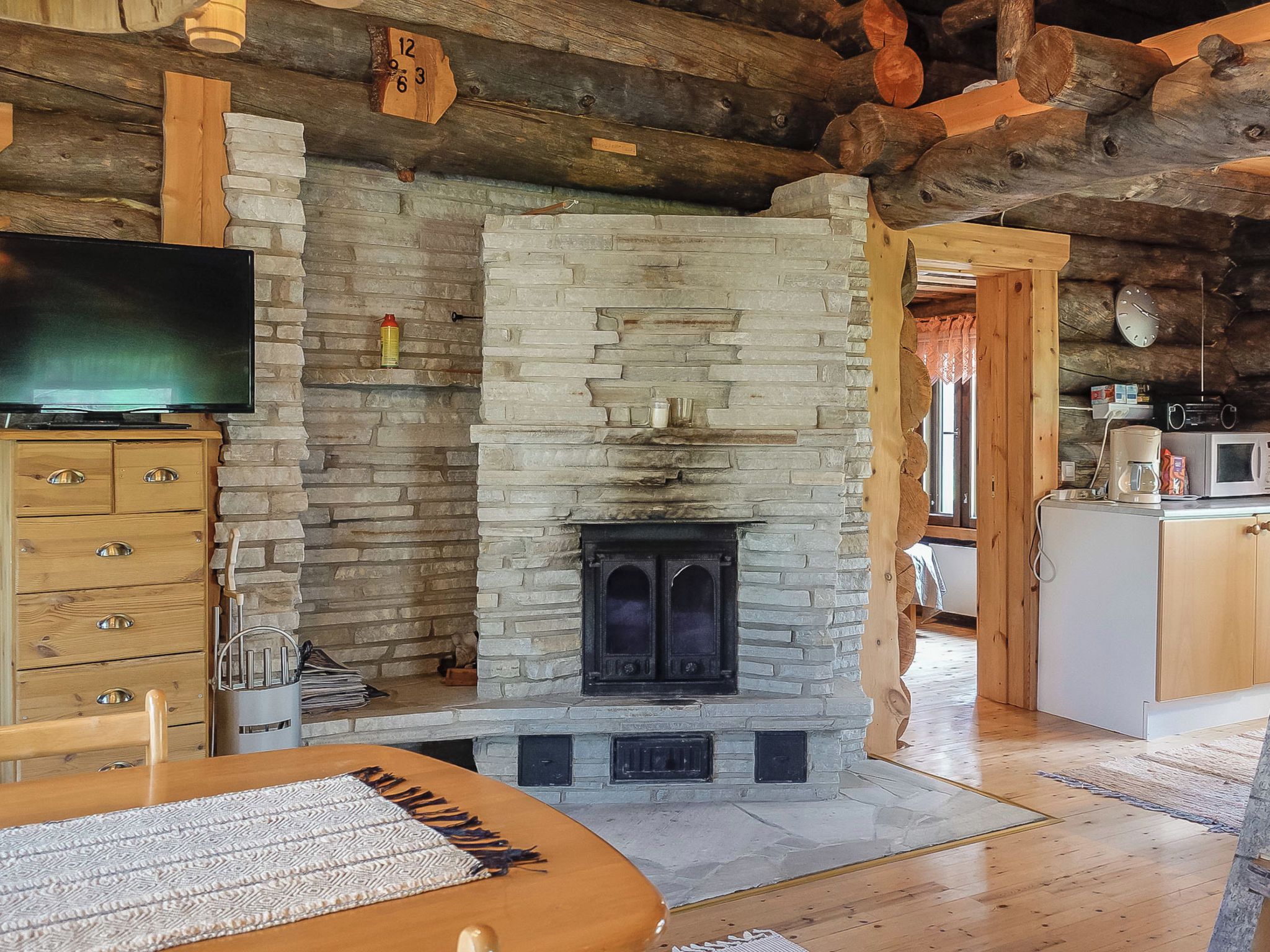 A cozy interior with a stone fireplace, wooden beams, a television, and a kitchen area.