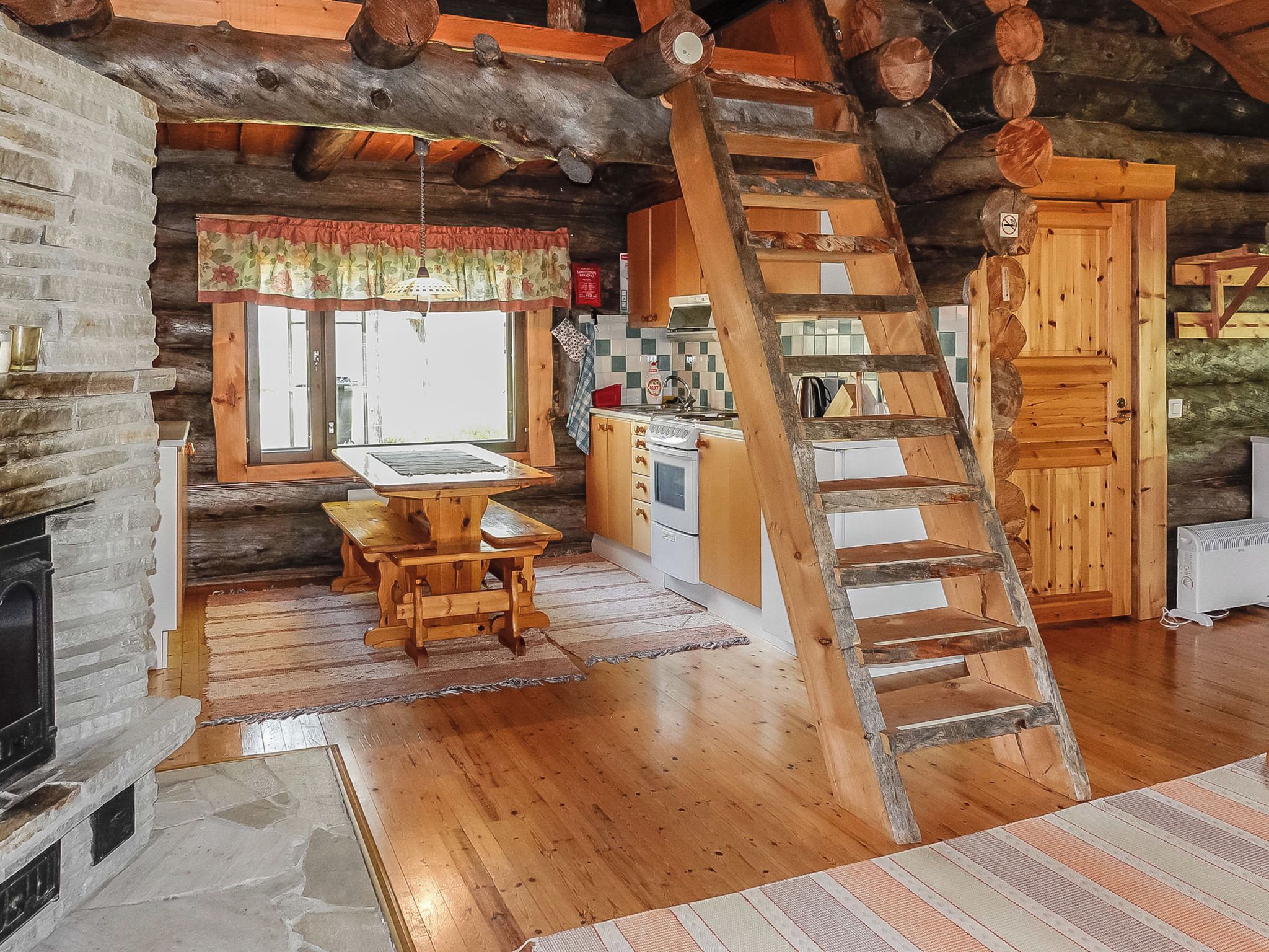 A rustic cabin interior with a wooden dining table, a kitchen area, and a wooden ladder leading to a loft.