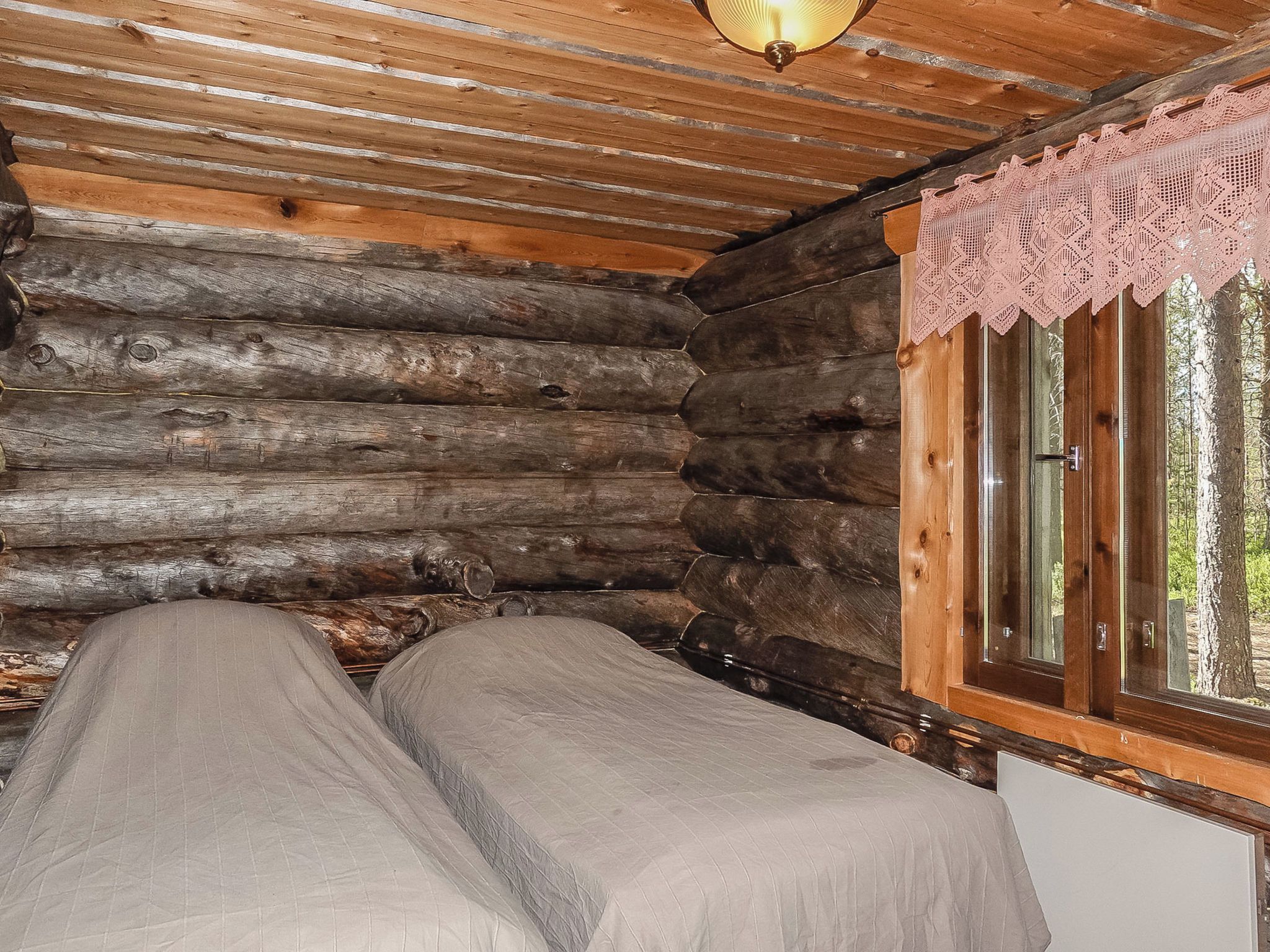 A log cabin room with a bed, a window with a lace curtain, and a wooden ceiling.