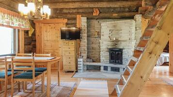 A cozy cabin interior with a wooden dining table, chairs, a TV, a stone fireplace, and a wooden staircase.