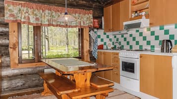 A rustic cabin kitchen with a wooden table and benches, a stove, and a view of the forest through large windows.
