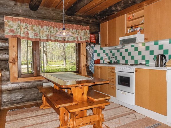 A rustic cabin kitchen with a wooden table and benches, a stove, and a view of the forest through large windows.