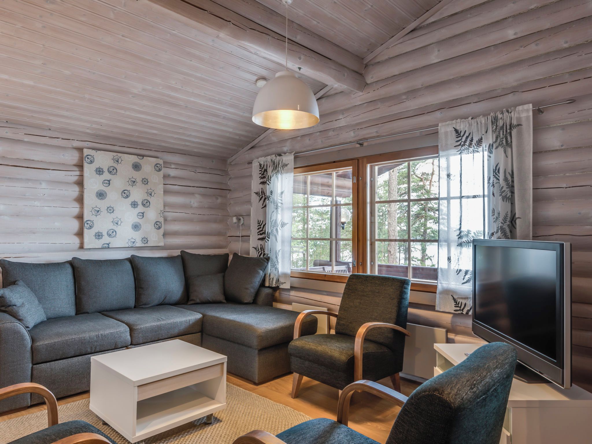 Un intérieur de chalet chaleureux avec un canapé d’angle, un fauteuil, un téléviseur et une grande fenêtre agrémentée de rideaux.