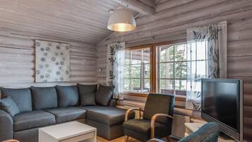 Un intérieur de chalet chaleureux avec un canapé d’angle, un fauteuil, un téléviseur et une grande fenêtre agrémentée de rideaux.