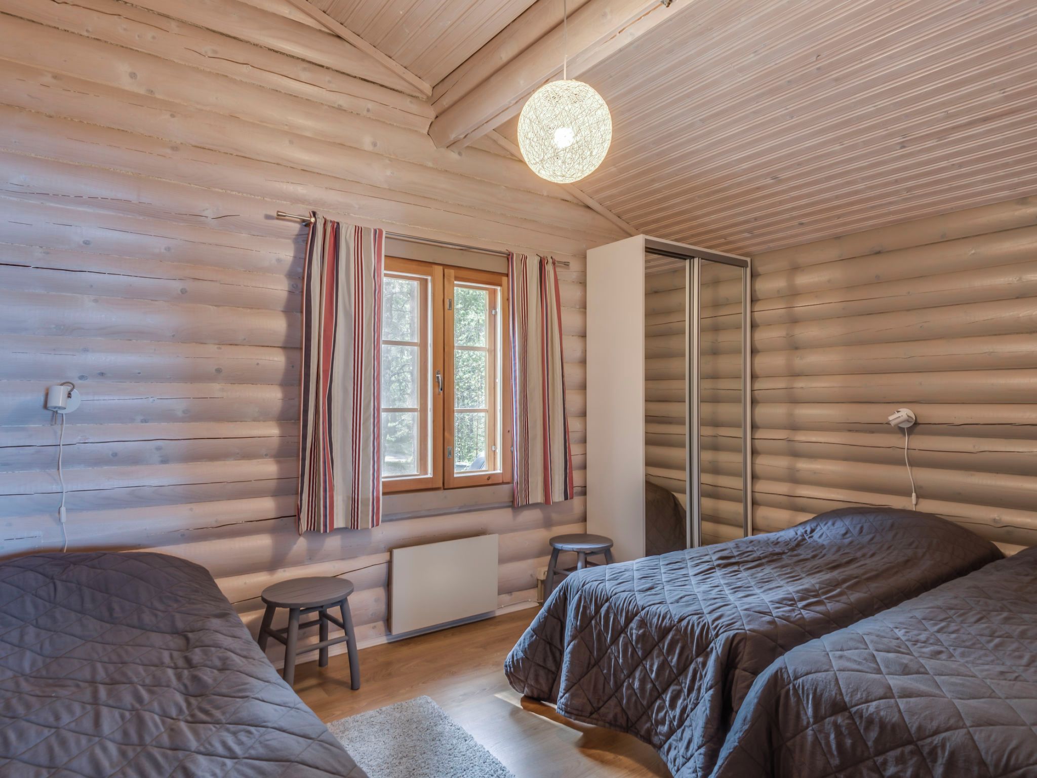 A log cabin room with two beds, a window with curtains, a radiator, and a hanging light fixture.