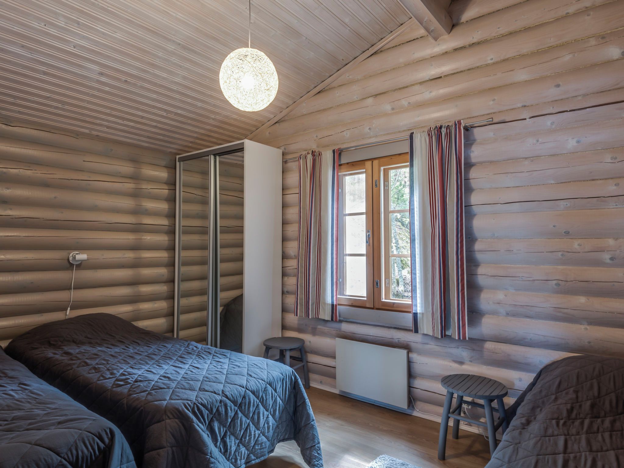 Een blokhutkamer met twee bedden, een raam met gordijnen, een radiator en een hanglamp.