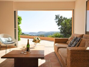 Uma sala de estar moderna com uma janela grande, um sofá de vime, uma poltrona e uma mesa com uma garrafa e copos.