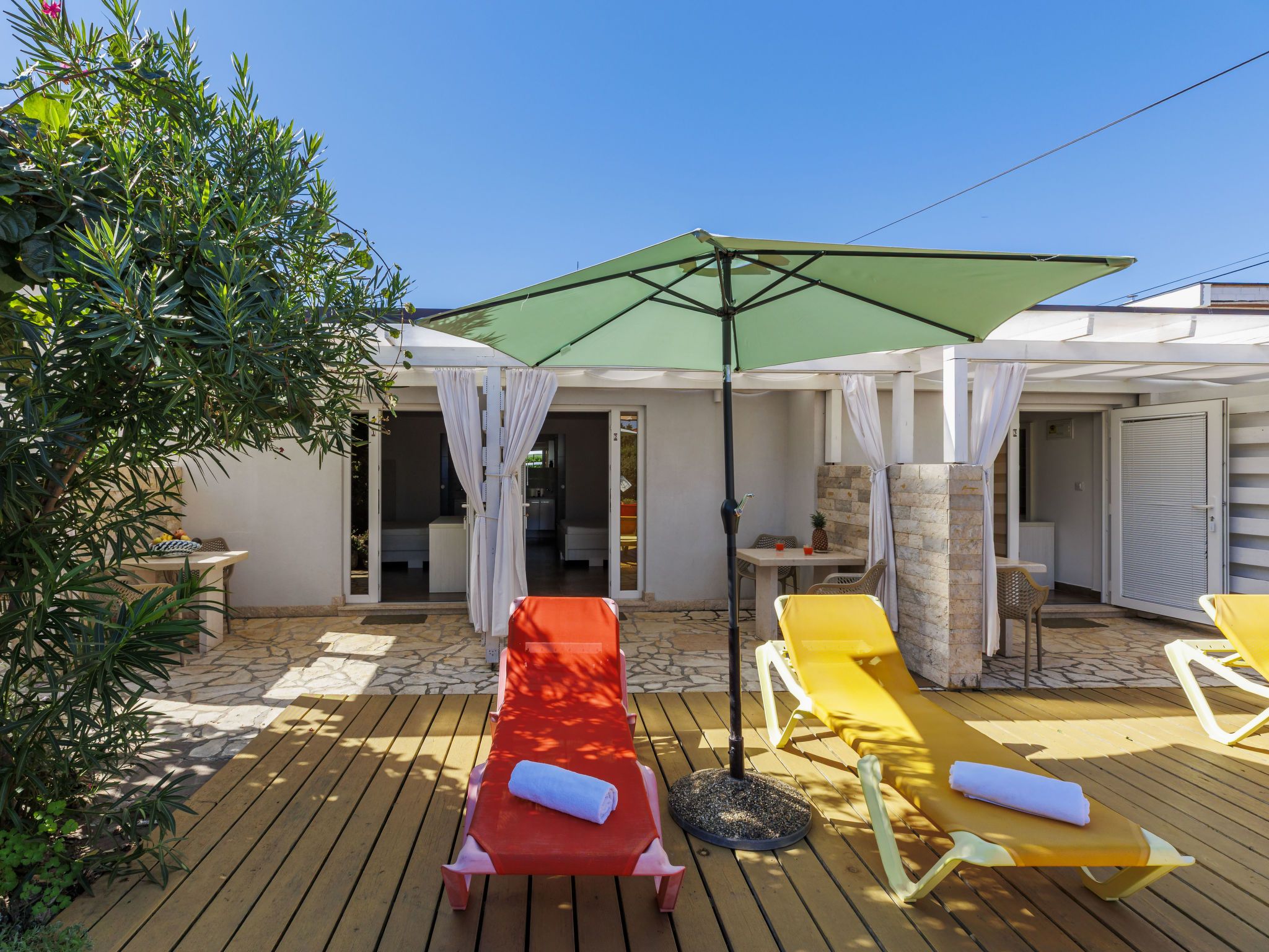 A wooden deck with sun loungers and a green umbrella, surrounded by a white building with stone walls.