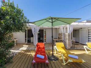 A wooden deck with sun loungers and a green umbrella, surrounded by a white building with stone walls.