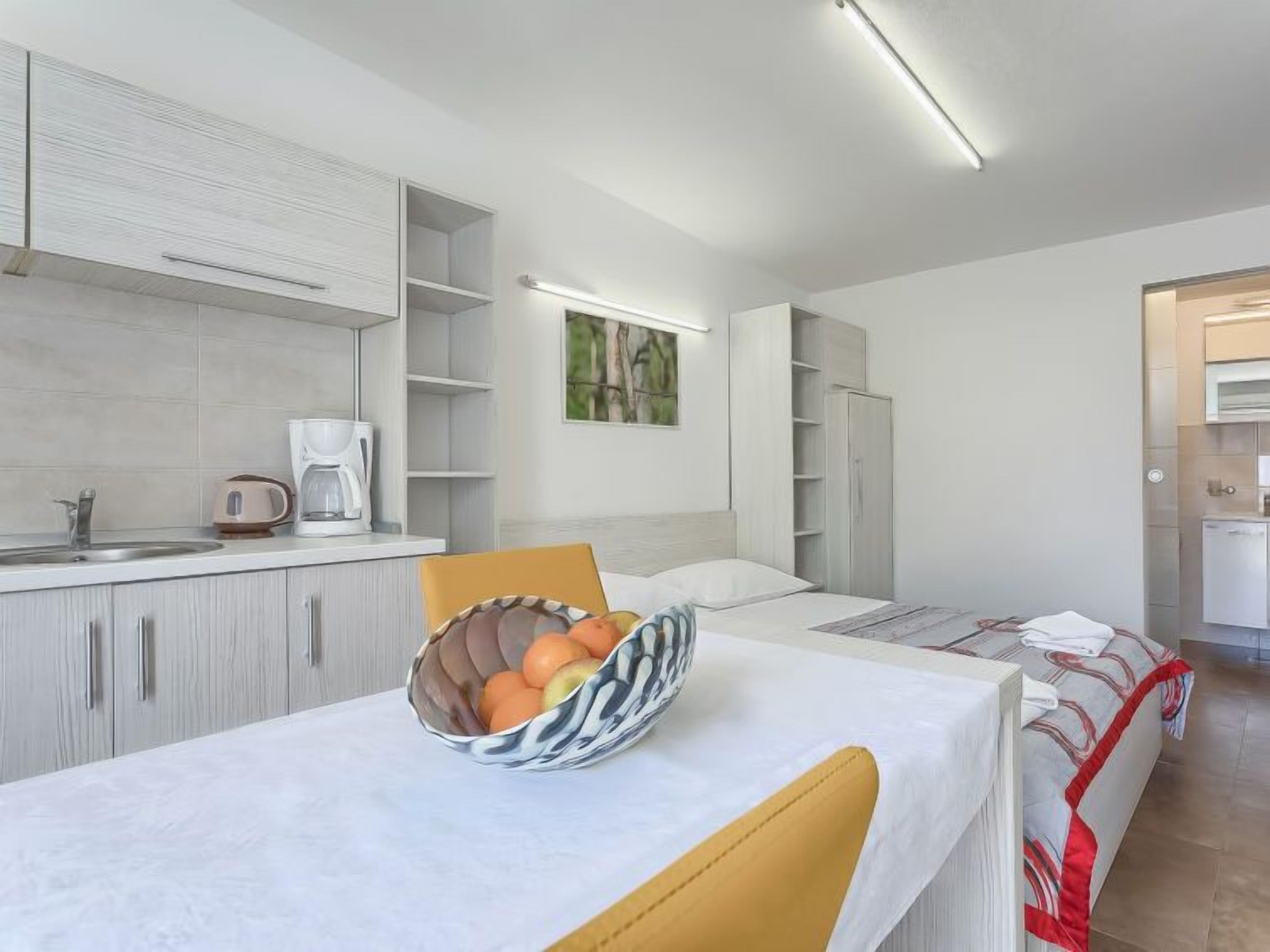 Une cuisine moderne avec des meubles blancs, une table à manger sur laquelle se trouve un saladier rempli de fruits, et un lit recouvert d’une couverture rouge et blanche.