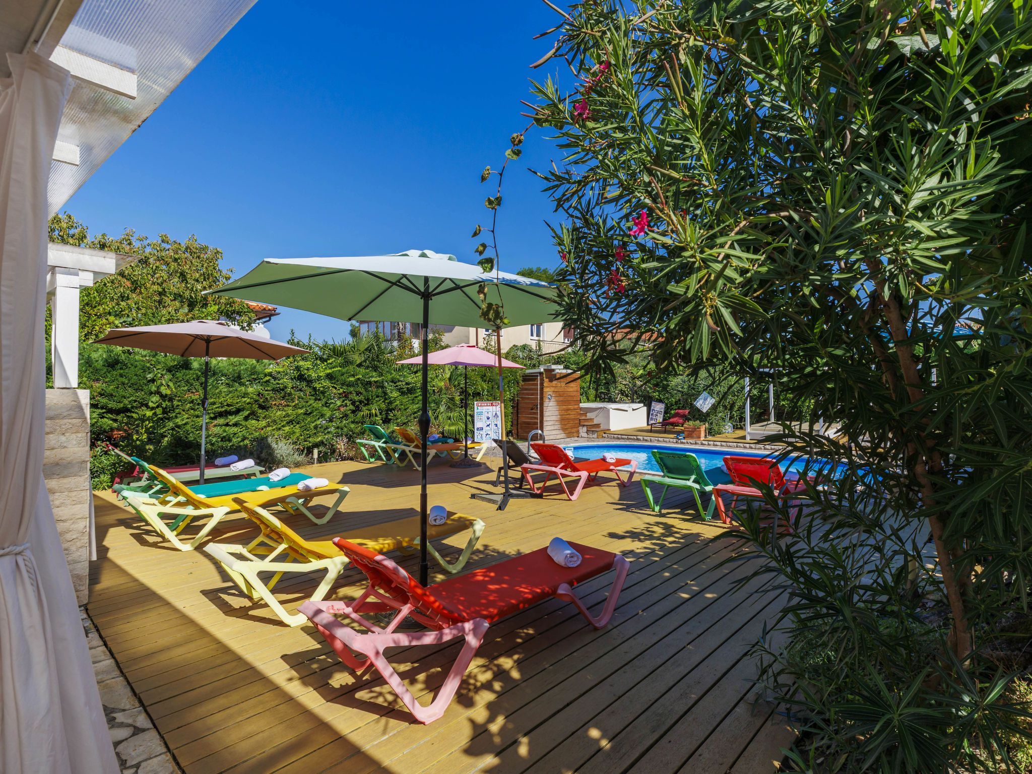 A wooden deck with colorful lounge chairs and umbrellas, a swimming pool, and lush greenery.
