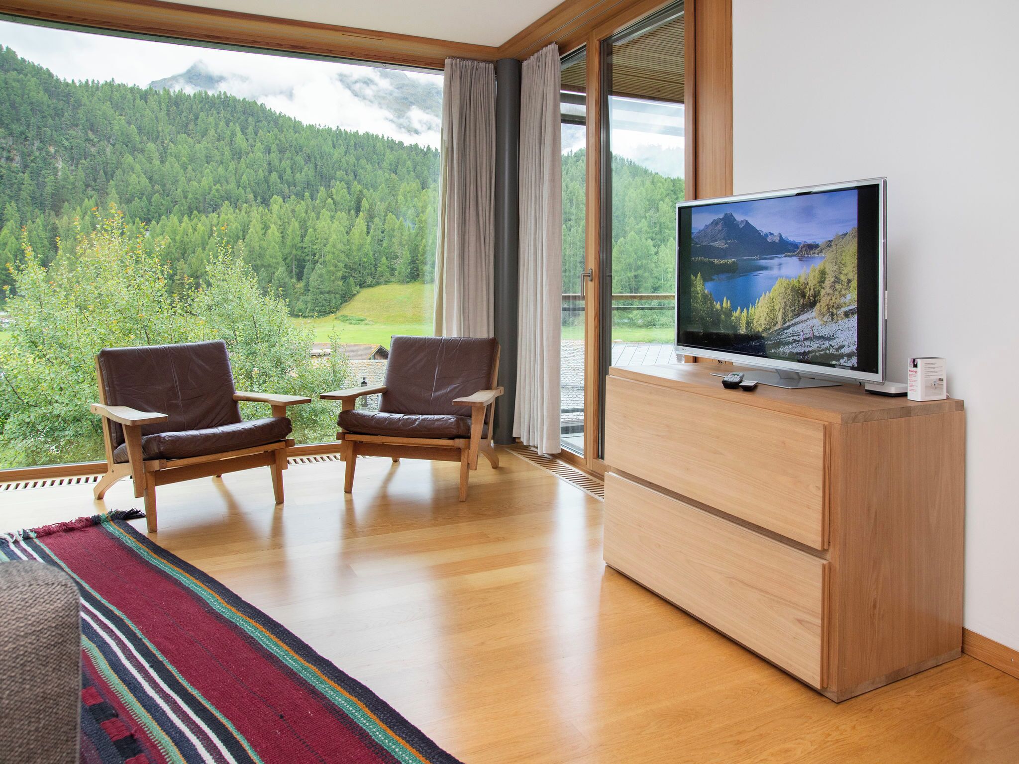 Ein Zimmer mit großem Fenster, zwei Ledersesseln, einer Holzschrank und einem Flachbildfernseher, der eine schöne Landschaft zeigt.