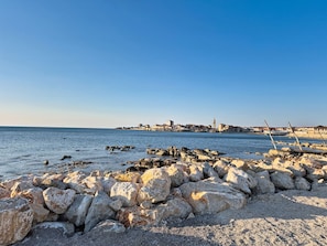 Paisaje costero con costa rocosa, mar tranquilo y un pueblo lejano con edificios y una torre destacada.