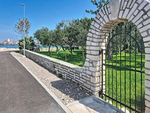 A paved walkway with a stone wall and metal gate, leading to a grassy area with trees.