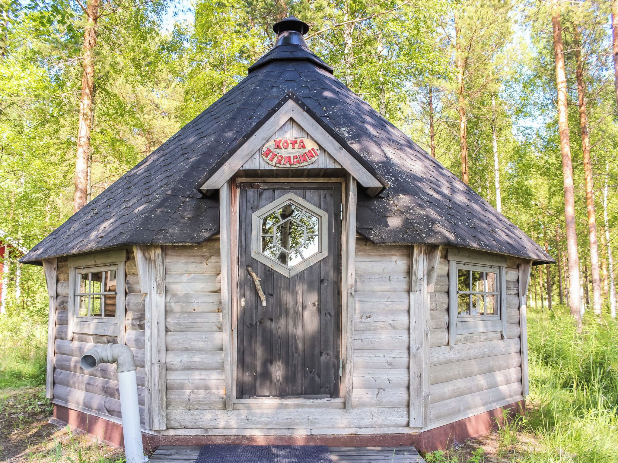 Puuinen mökki, jossa on kuusikulmainen ikkuna, savupiippu ja kyltti, jossa lukee ”Kota Hermann”.