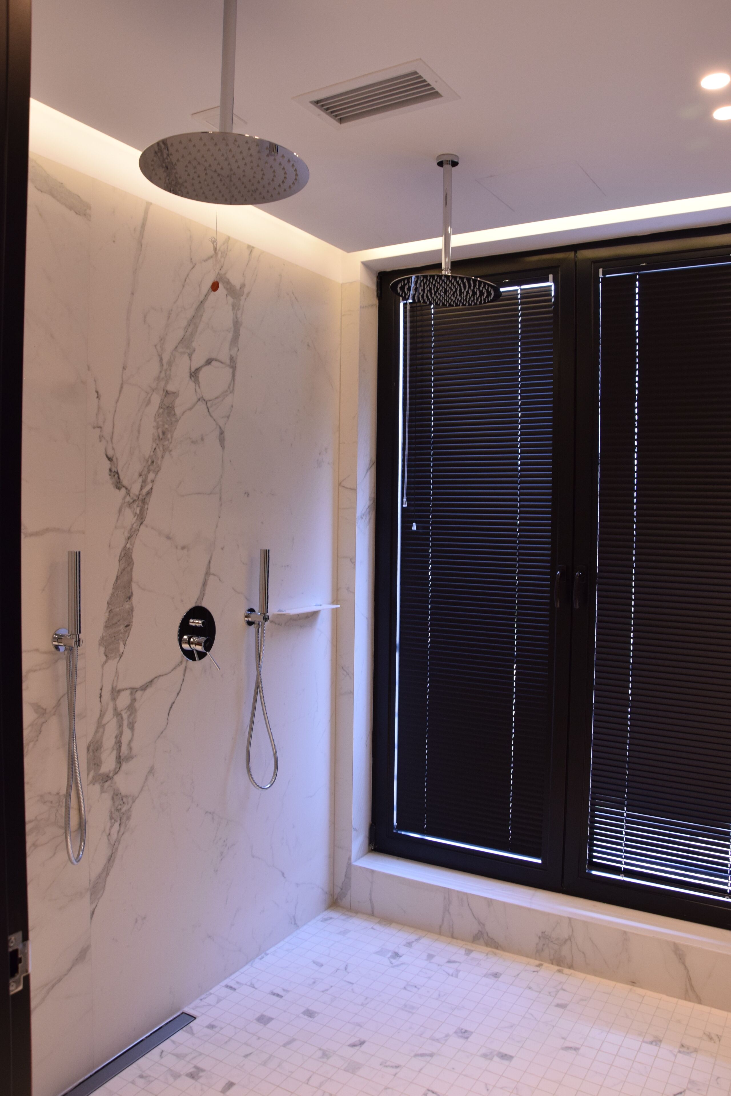 A modern bathroom with a marble wall, a large window with blinds, and a shower with a circular light fixture.