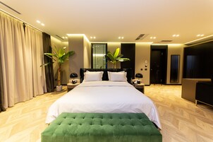 Un dormitorio de hotel moderno con una cama grande, dos mesitas de noche, un otoman verde y una planta en maceta.