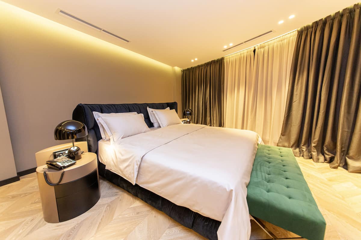 A modern hotel room with a large bed, a bedside table with a lamp, and a green bench.