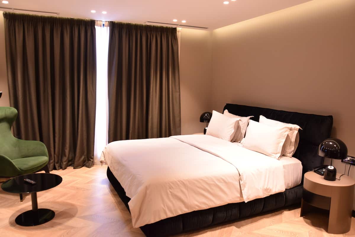A modern hotel room with a large bed, a green armchair, a small round table, and a bedside table with a lamp.