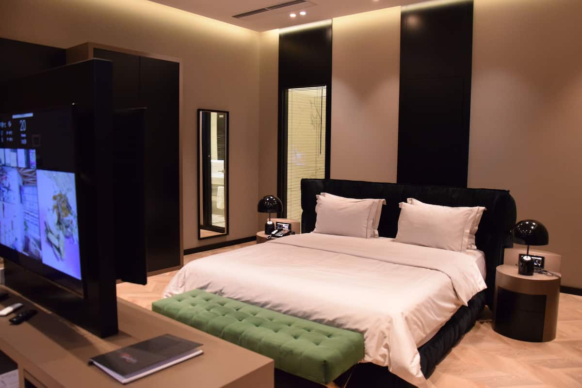 A modern hotel room with a large bed, a flat-screen TV, a desk with a laptop, and a green ottoman.