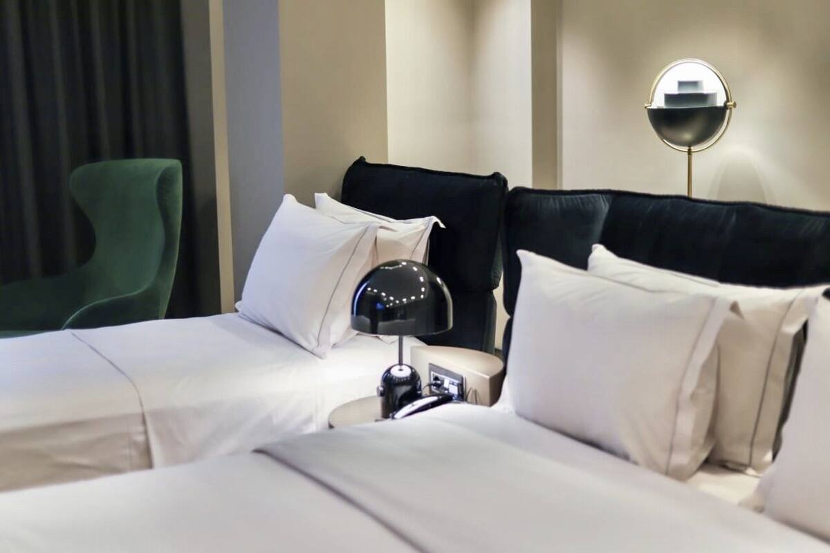 A hotel room with a neatly made bed, a green armchair, and a bedside table with a lamp.