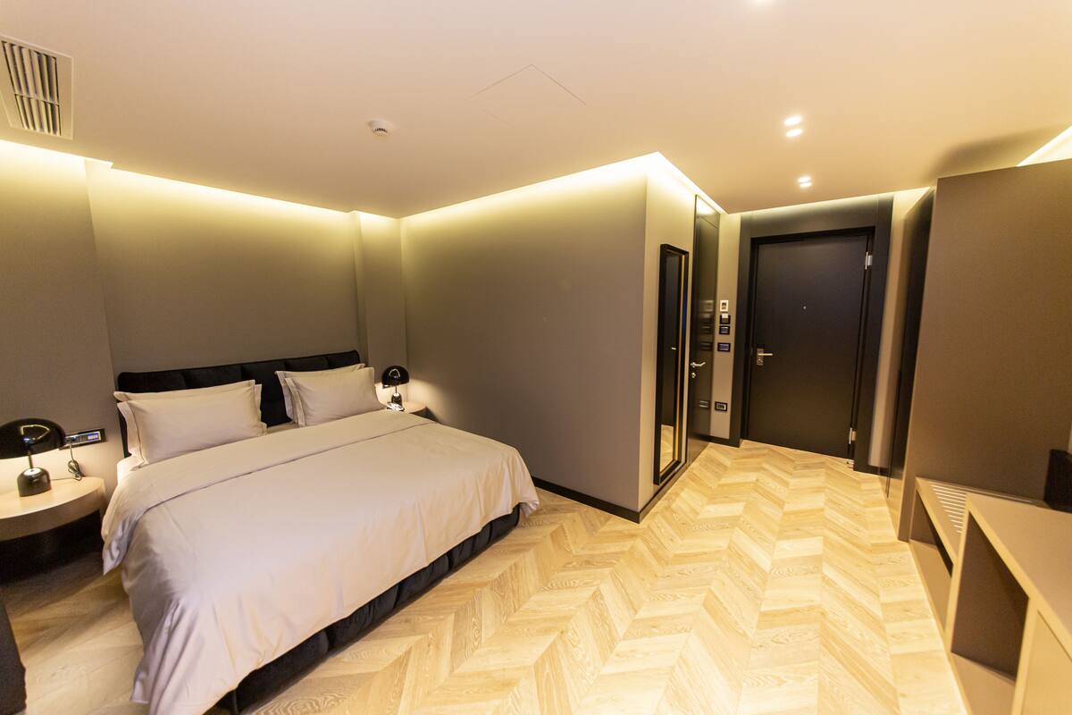 A modern hotel room with a large bed, bedside tables, a wardrobe, and a herringbone-patterned floor.