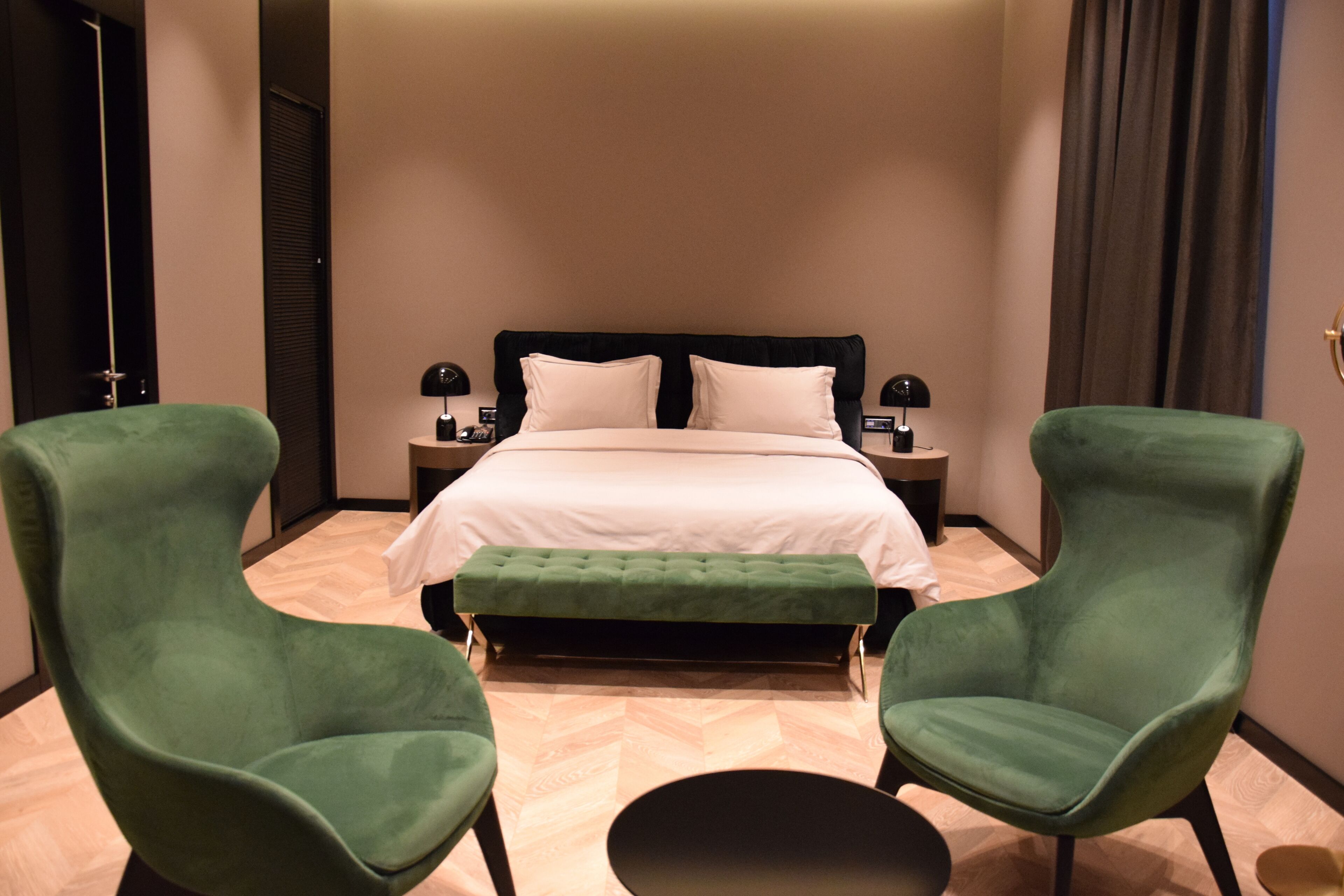Una habitación de hotel con una cama, dos sillones verdes, una mesa redonda y una mesita de noche con lámpara.