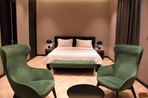 Una habitación de hotel con una cama, dos sillones verdes, una mesa redonda y una mesita de noche con lámpara.