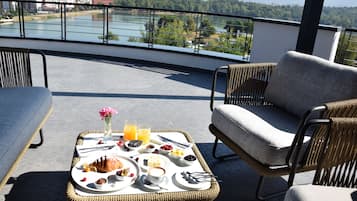 Varanda com mobiliário de vime, bandeja de café da manhã com croissants, frutas e bebidas, e vista para a cidade e rio.