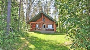 Une cabane avec un porche à l’avant, une petite terrasse et une cour gazonnée entourée d’arbres.