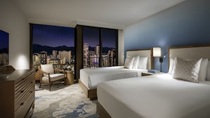 Ein Hotelzimmer mit zwei Betten, einem Schreibtisch, einem Stuhl und einer Kommode. Das Zimmer verfügt über ein großes Fenster mit Blick auf die Stadt.