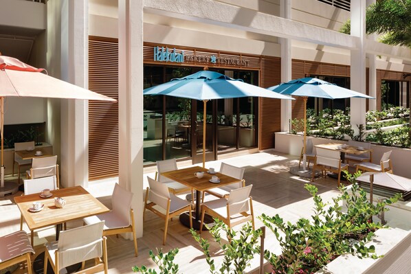 An outdoor cafe area with wooden tables and chairs, blue umbrellas, and a view of a restaurant named "Halekulani". - Halepuna Waikiki by Halekulani (Honolulu)