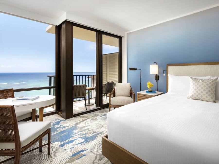A hotel room with a large bed, a desk, a chair, and a balcony with a view of the ocean.