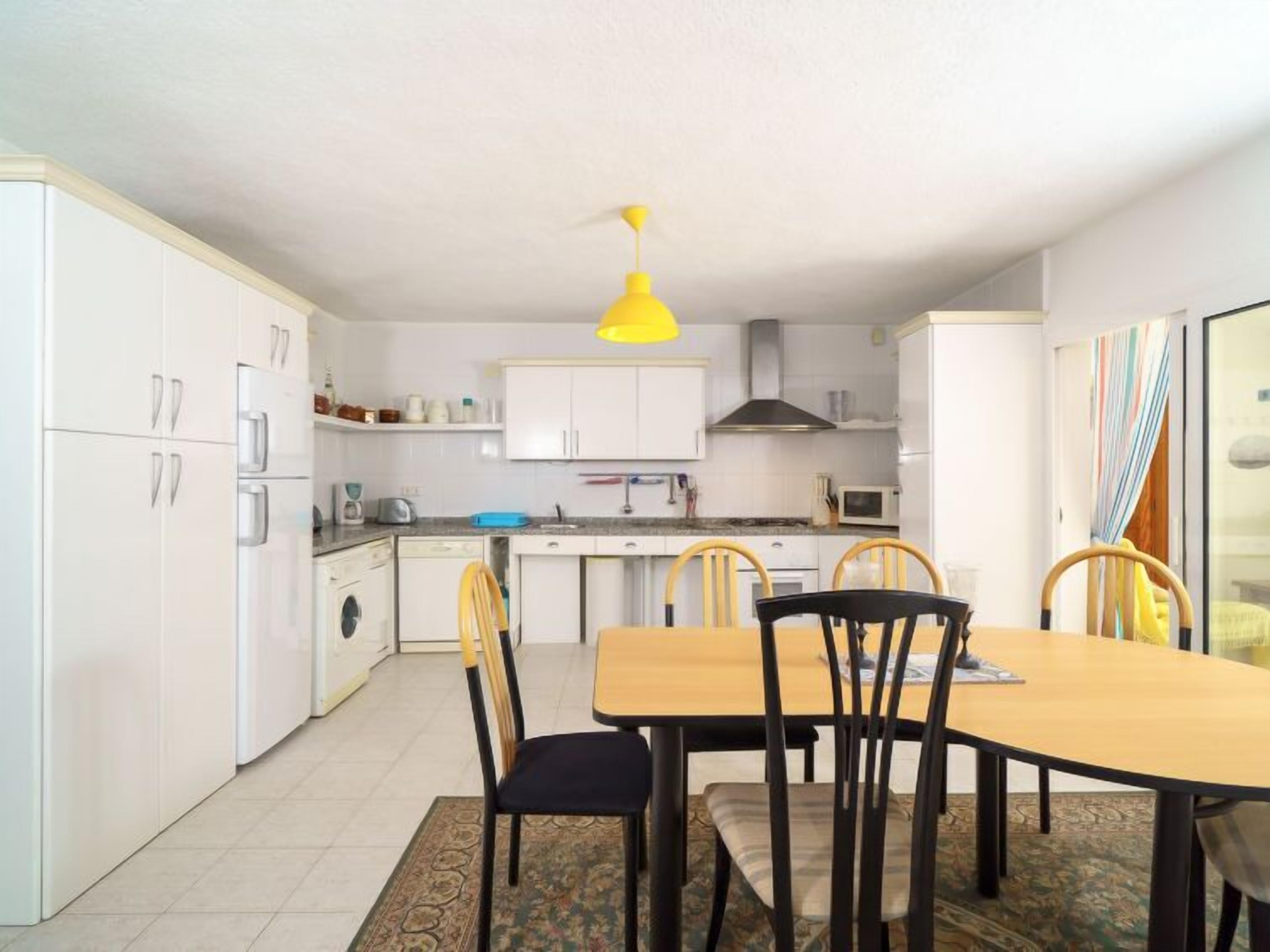 Una cocina compacta con armarios blancos, una lámpara colgante amarilla, una mesa de comedor con cuatro sillas y un tapete estampado.