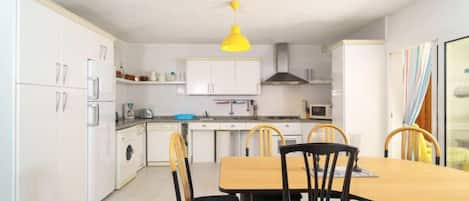 Una cocina compacta con armarios blancos, una lámpara colgante amarilla, una mesa de comedor con cuatro sillas y un tapete estampado.